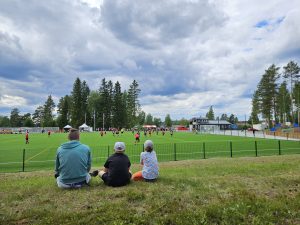 Aikuinen ja kaksi lasta istuvat nurmikolla ja seuraavat jalkapallo-ottelua.