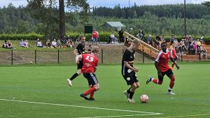 Jalkapallo-ottelusta pelitilanne, mustapaitainen HaKoPo:n pelaaja pitää palloa, kaksi punapaitaista vastustajaa yrittää saada pallon itselleen. Taustalla yleisöä istumassa katsomossa ja seuraamassa peliä.