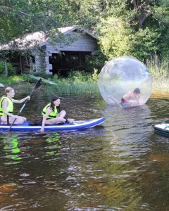 Lapset harrastavat vesiaktiviteetteja kesällä, kaksi lasta melomassa pelastusliivit päällä SUP-laudalla, yksi lapsi on suuren Zorb-vesipallon sisällä. Aurinko paistaa taustan puihin, rannalla näkyy hirsinen rakennus.