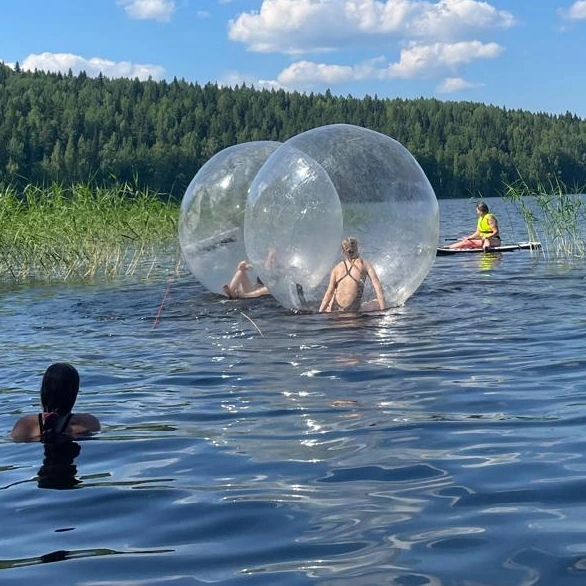 Kesäinen järvi, kaukana vastarannalla vihreä metsä. Yksi lapsi on uimassa, kaksi istuu Zorb-vesipallojen sisällä ja taustalla yksi meloo SUP-laudalla.