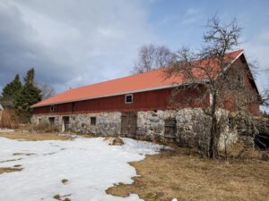 Iso, vanha navettarakennus. Alaosa kiviä, yläosa punaiseksi maalattua lankkua, punainen uusi katto. Maassa vielä vähän lunta.