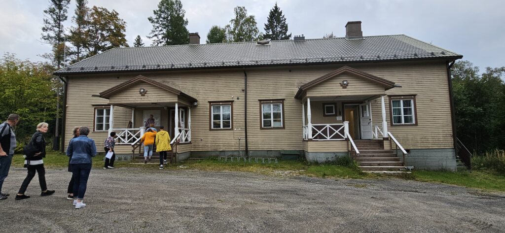 Suuri ruskea puutalo. Sisään vie kaksi ovea, joissa molemmissa portaikot. Neljä ikkunaa. Ihmisiä menossa vasemmanpuoleisesta ovesta sisään.