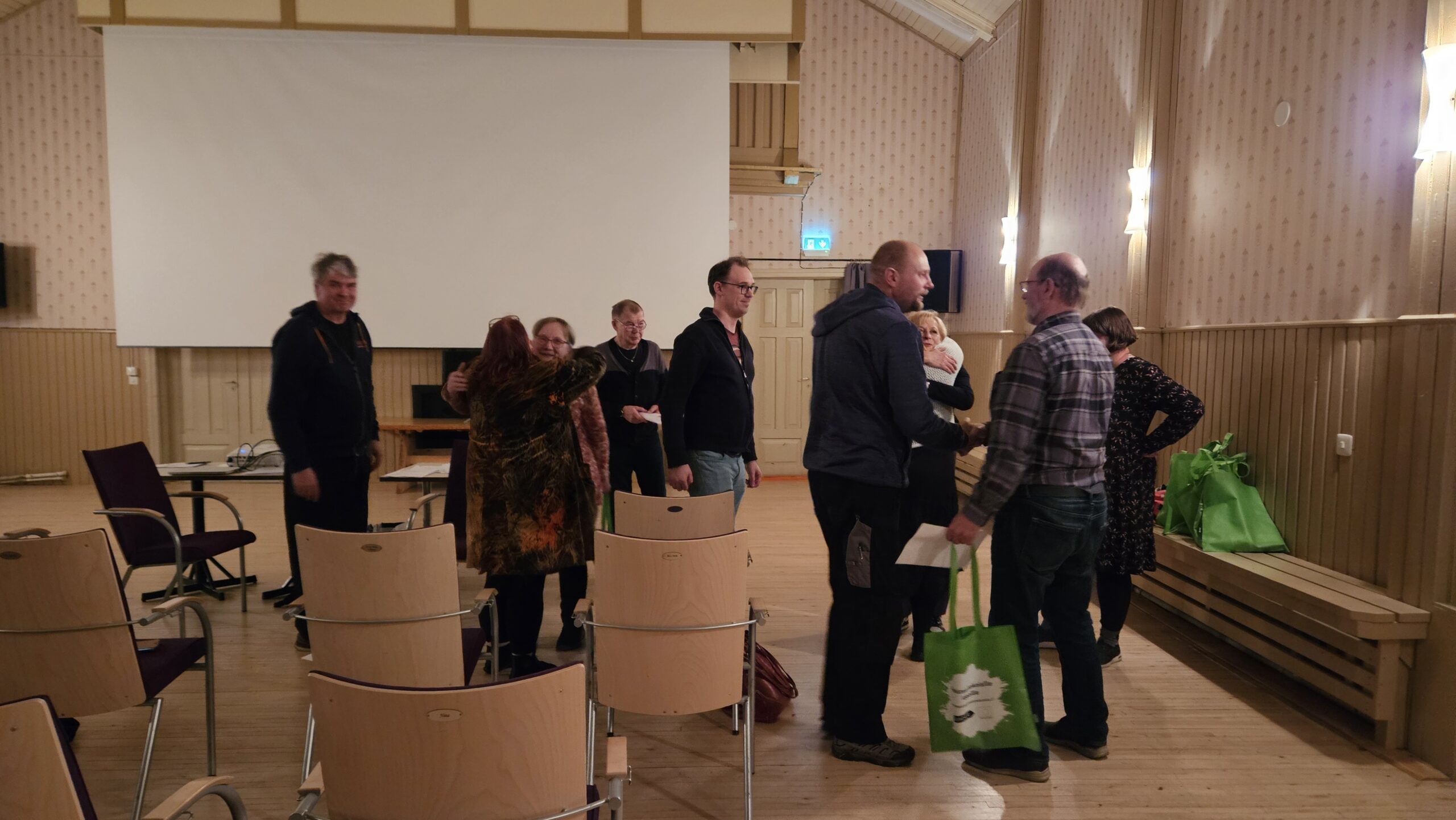 Suuri sali, jonka taustalla valkokangas, etualalla tuoleja. Keskellä kymmenen ihmistä, jotka sanovat jäähyväisiä toisilleen. Kaksi ihmistä kättelee ja neljä on halaamassa, loput odottavat vuoroaan.
