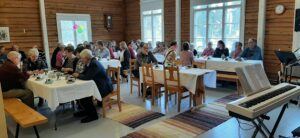 Tila, jossa hirsiseinät. Lattioilla räsymattoja, pöydissä valkoiset pöytäliinat ja ihmiset istuvat pöytien ympärillä kahvittelemassa.