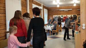 Hirsiseinäinen huone, jonka takaseinällä on käsitöitä nostettu ilmoitustaululle näyttelyyn. Ihmisiä jonottamassa kahville vasemmalla seinustalla. Osa istuu jo pöytien ympärillä huoneen takaosassa.