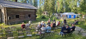 Yleisöä istumassa tuoleilla ulkona vanhan hirsituvan vieressä, he kuuntelevat konserttia. Taustalla näkyy telttoja.