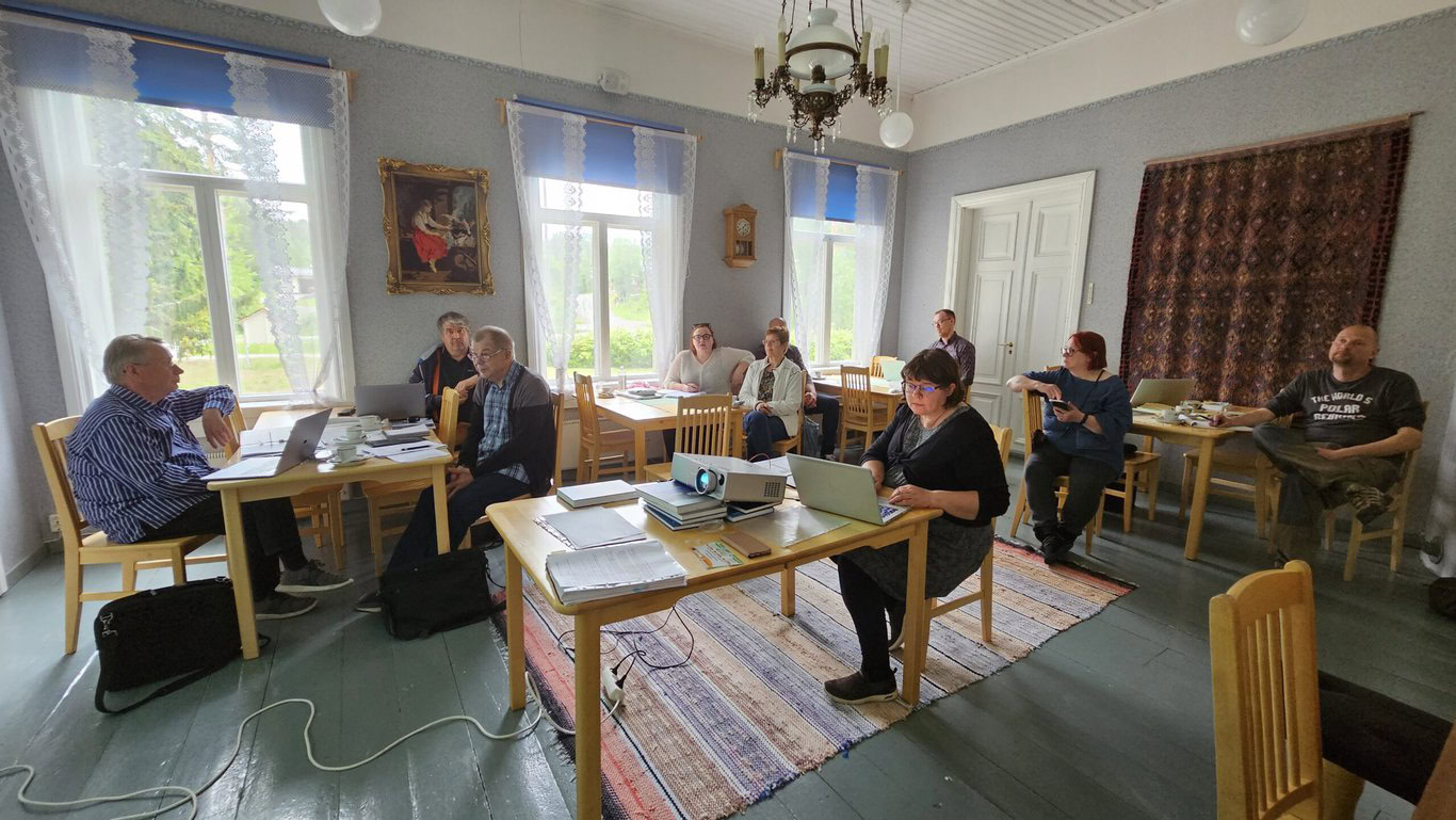 Kymmenen henkilöä istuu viiden eri pöydän ääressä kokoustamassa. Useammalla on kannettava tietokone avoinna, yhdellä pöydällä on käynnissä oleva videotykki. Huoneessa on taustalla kolme ikkunaa ja toisella seinällä ovi, jonka vieressä iso ryijy.