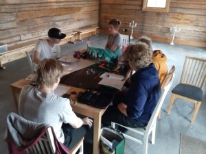 Viisi nuorta henkilöä on kokoontunut pöydän ympärille pelaamaan. 