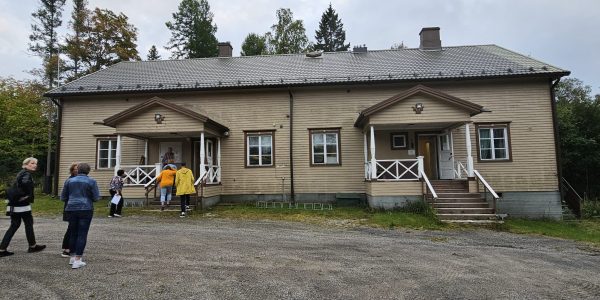 Suuri ruskea puutalo. Sisään vie kaksi ovea, joissa molemmissa portaikot. Neljä ikkunaa. Ihmisiä menossa vasemmanpuoleisesta ovesta sisään.