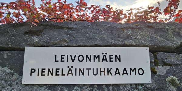 Kyltissä yrityksen nimi Leivonmäen pieneläintuhkaamo. Kyltti kiinnitetty kellarin kiviseinään, ylhäällä punaista pensaskasvustoa.